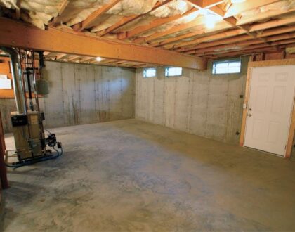 An unfinished basement with exposed insulation in the ceiling and concrete walls. The floor appears bare, and there is a furnace and utility meter installed on the left, with a white door on the right providing entry or exit.